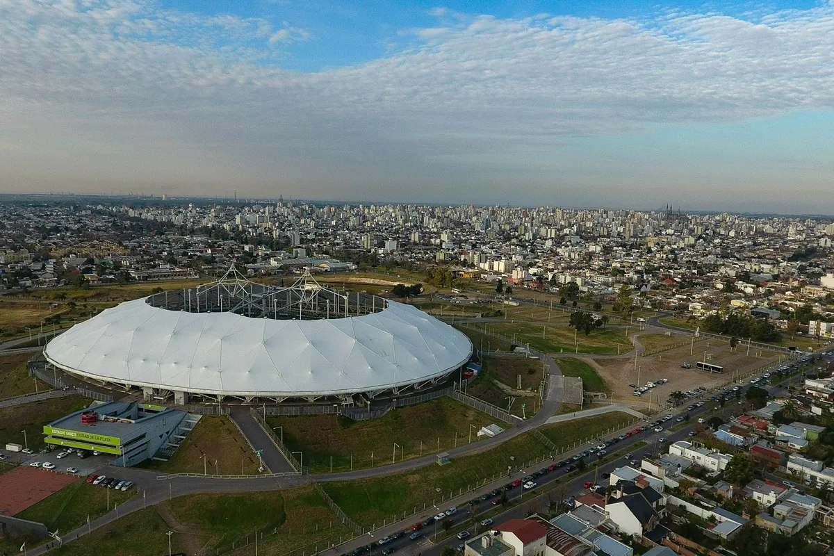 Vista aérea del Estadio Único de La Plata: un estadio ovalado con techo blanco inclinado, mástiles y cables de sostén, rodeado de parques, accesos, estacionamientos y edificios de la ciudad, bajo un cielo azul con bandas de nubes.