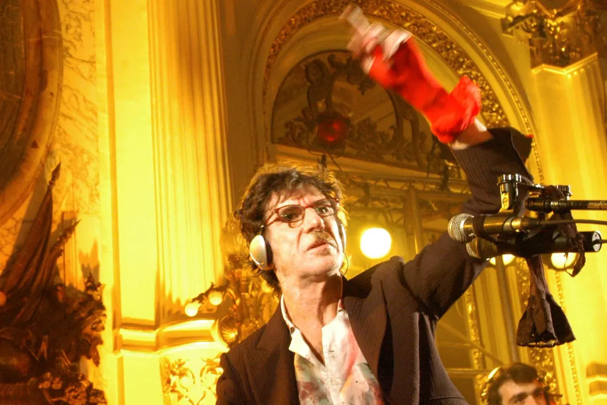 Charly García, con anteojos, auriculares y saco oscuro sobre una camisa blanca manchada de pintura, levanta un brazo con un guante rojo en un salón dorado y ornamentado, frente a varios micrófonos.