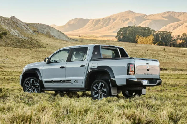 Una Volkswagen Amarok V6 gris clara, vista de tres cuartos desde atrás, en un prado con rocas y montañas iluminadas por una luz dorada al fondo.