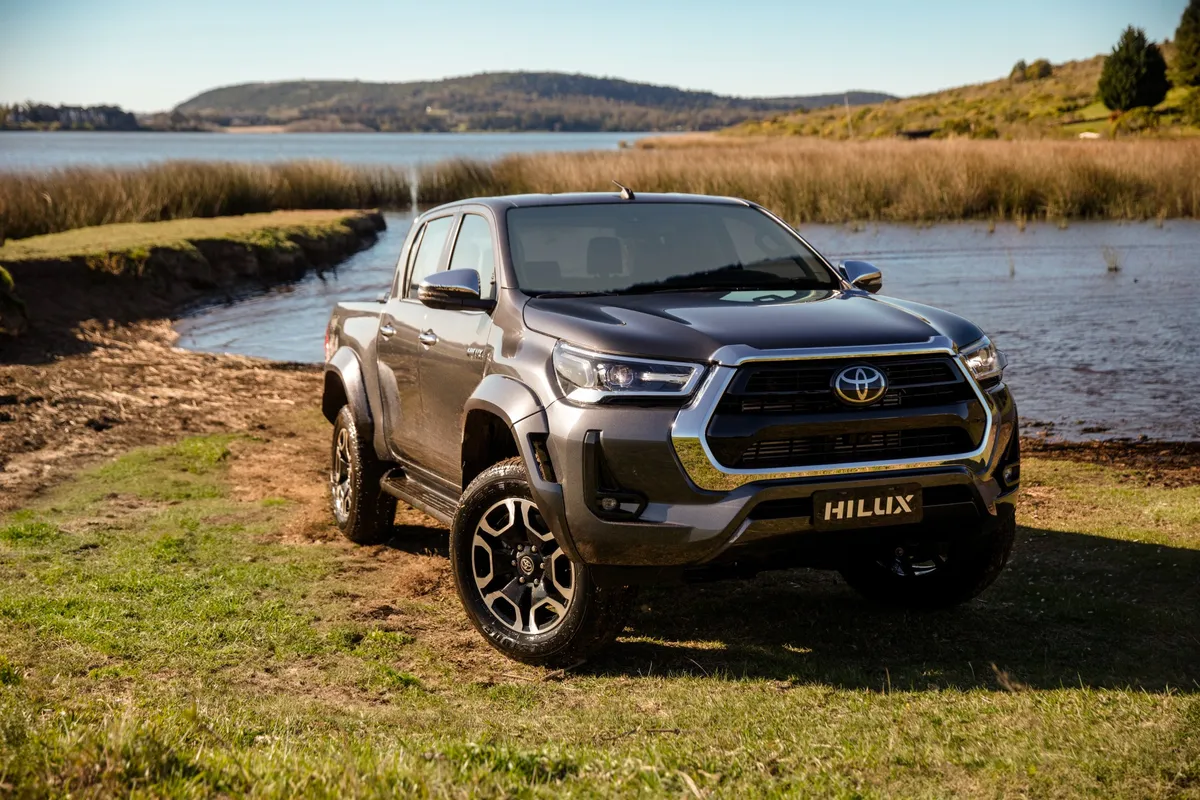 Una Toyota Hilux gris metalizada, vista de tres cuartos de frente, estacionada sobre una orilla con pasto junto a un lago rodeado de juncos y colinas, bajo un cielo despejado.