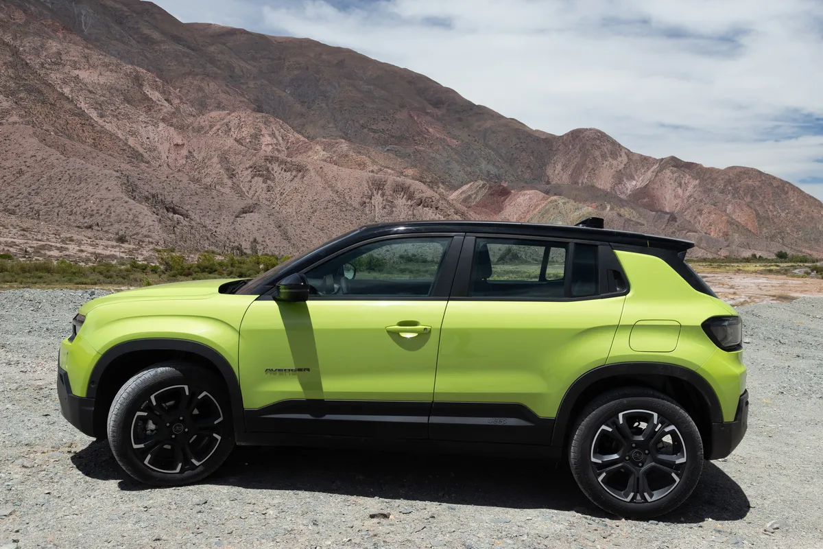 Un Jeep Avenger verde con techo negro, de perfil, sobre ripio frente a laderas rocosas.