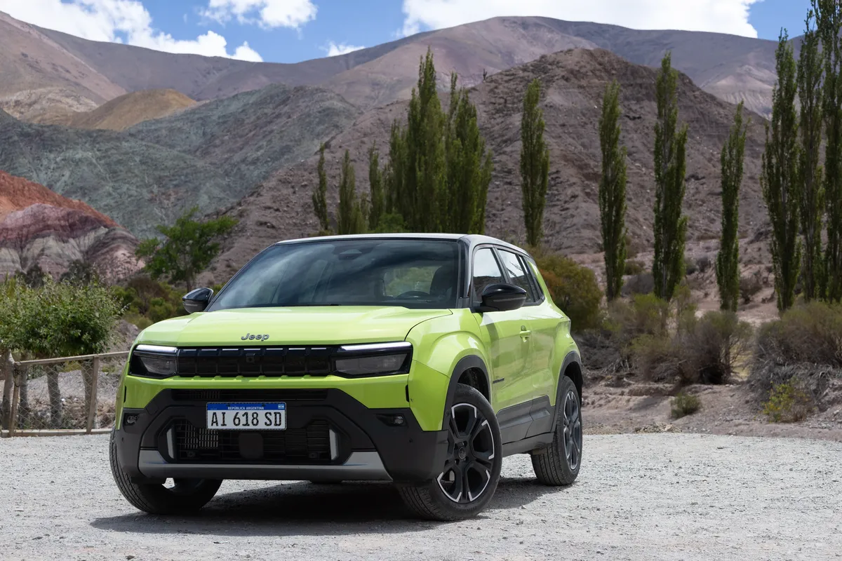 Un Jeep Avenger verde casi de frente, en un claro de ripio con árboles, un alambrado y montañas.