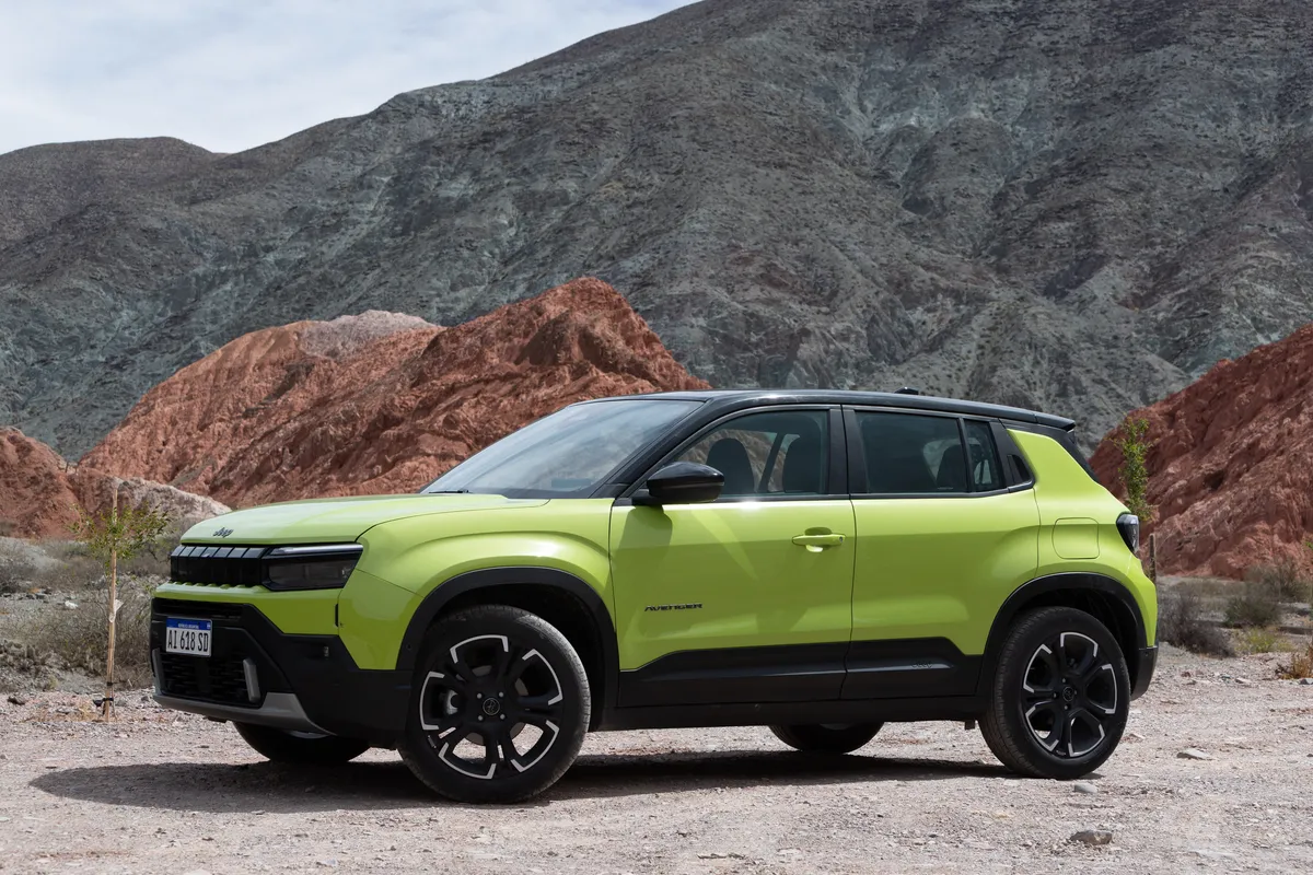 Un Jeep Avenger verde casi de lado, sobre ripio, con montañas de varios colores al fondo.