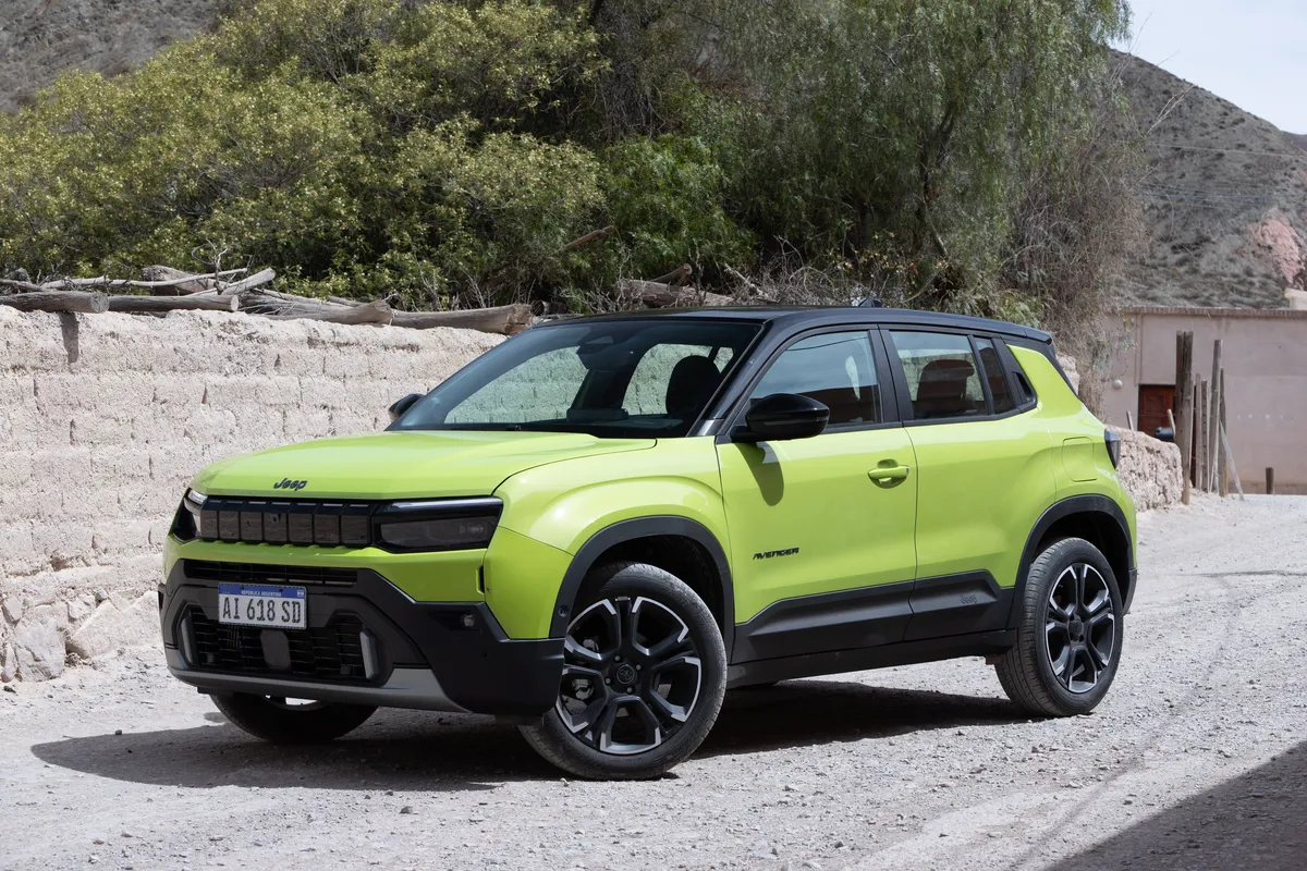 Un Jeep Avenger verde de tres cuartos de frente, estacionado junto a una pared de adobe y árboles.