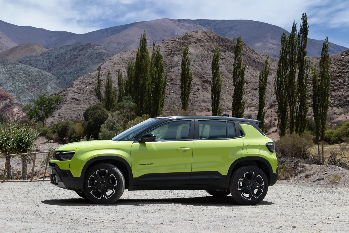 Un Jeep Avenger verde de perfil, junto a un alambrado rústico, árboles y montañas.