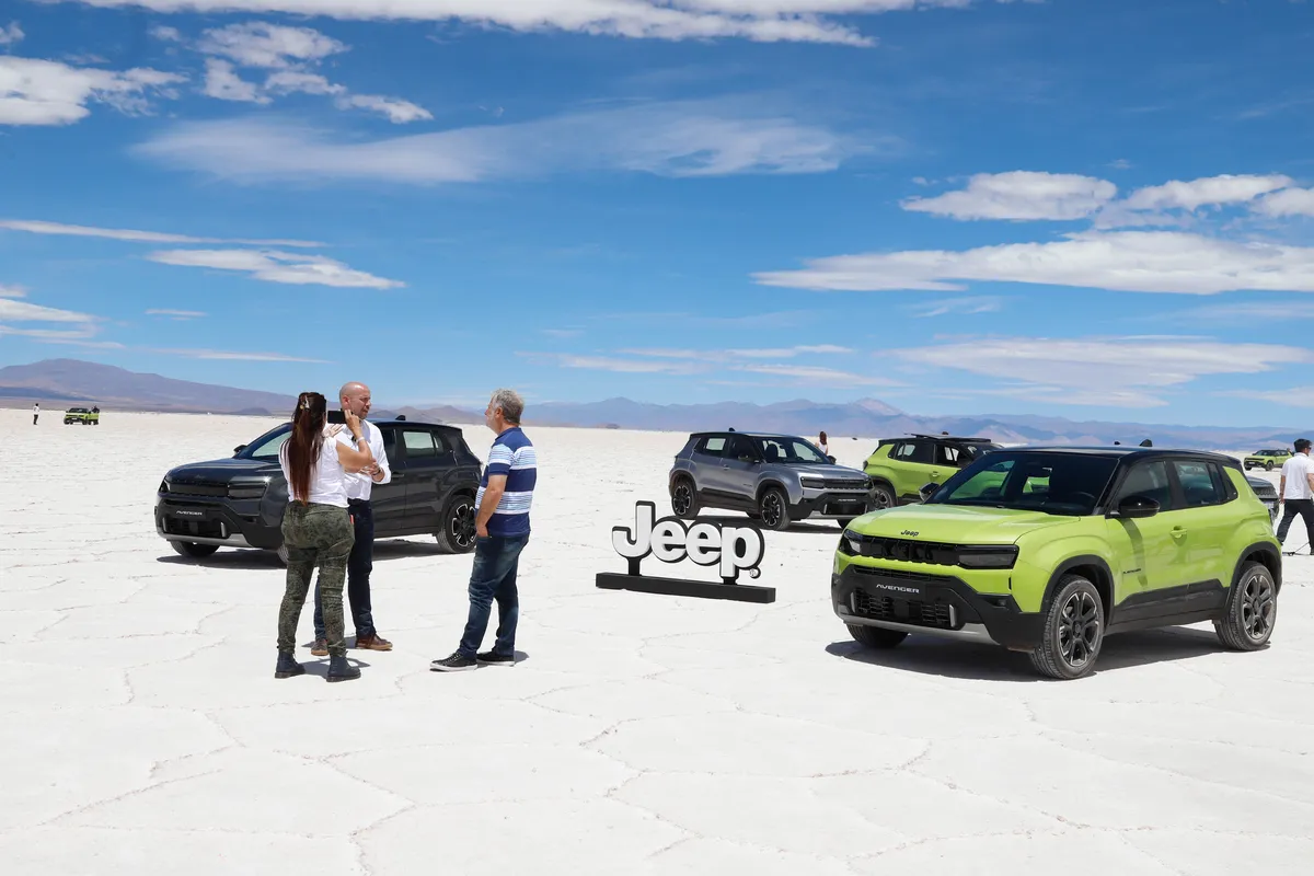 Un Jeep Avenger verde de tres cuartos de frente sobre un salar blanco, con otras camionetas y personas detrás.