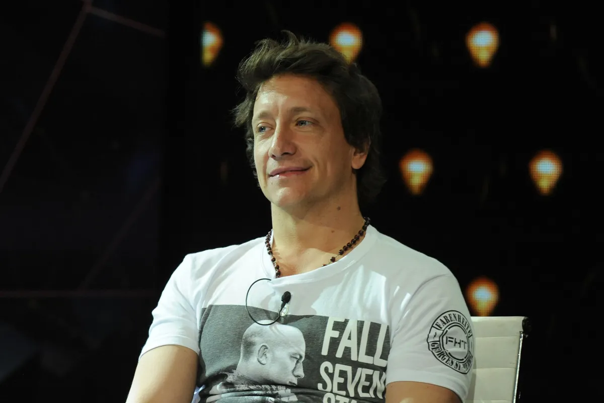 Nico Vázquez, sentado en un sillón blanco sobre un escenario oscuro, con remera blanca estampada y micrófono corbatero, sonríe mientras mira hacia un costado.