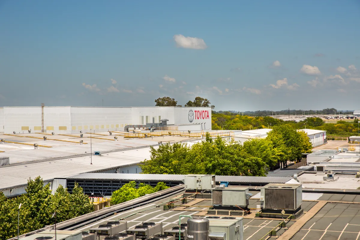 Vista elevada de los techos de la planta de Toyota en Zárate, con equipos de ventilación, árboles y un edificio blanco con el logo y el nombre TOYOTA en rojo bajo un cielo celeste.